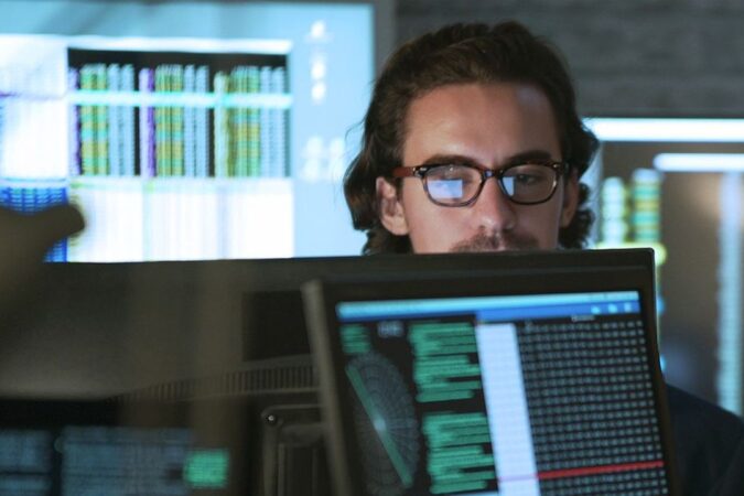 Stock photo of a young man & woman working in an office surrounded by computer monitors displaying moving numerical & scientific data.