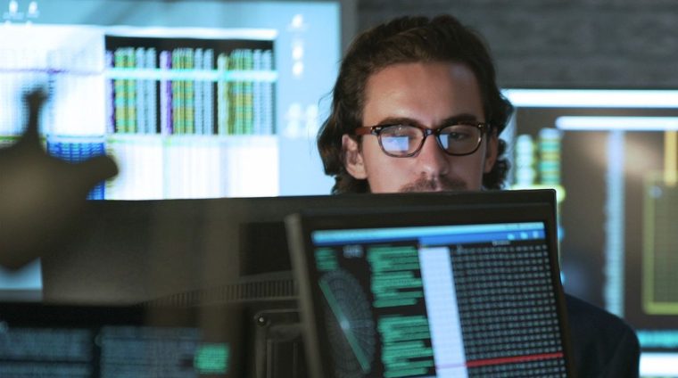 Stock photo of a young man & woman working in an office surrounded by computer monitors displaying moving numerical & scientific data.